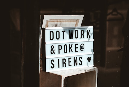 A light box sign with black text displaying the words 'DOT WORK & POKE @ SIRENS &hearts;' against a white background. The sign is placed on a wooden or metal surface, within a dimly lit interior environment.