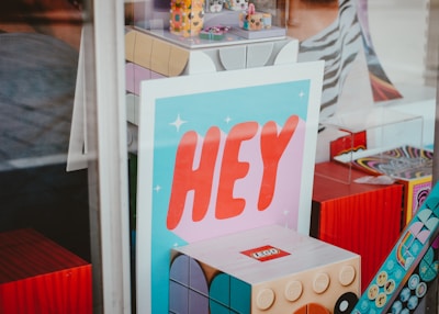 A window display featuring a prominent poster with the word 'HEY' in large red letters on a blue background. Surrounding the poster are various colorful and playful items, including a large LEGO-style brick and other geometric shapes with vibrant and whimsical designs.