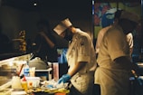 A dimly lit kitchen scene with chefs in white uniforms and hats preparing food. One of the chefs is wearing a black face mask and blue gloves while concentrating on the task at hand. The countertop is cluttered with various cooking supplies, utensils, and containers. In the background, a colorful partition with floral designs adds visual interest.