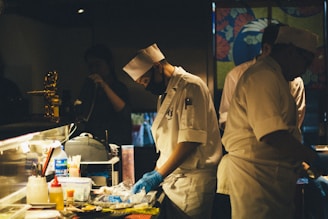 A dimly lit kitchen scene with chefs in white uniforms and hats preparing food. One of the chefs is wearing a black face mask and blue gloves while concentrating on the task at hand. The countertop is cluttered with various cooking supplies, utensils, and containers. In the background, a colorful partition with floral designs adds visual interest.