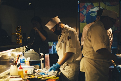 A dimly lit kitchen scene with chefs in white uniforms and hats preparing food. One of the chefs is wearing a black face mask and blue gloves while concentrating on the task at hand. The countertop is cluttered with various cooking supplies, utensils, and containers. In the background, a colorful partition with floral designs adds visual interest.