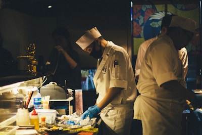 A dimly lit kitchen scene with chefs in white uniforms and hats preparing food. One of the chefs is wearing a black face mask and blue gloves while concentrating on the task at hand. The countertop is cluttered with various cooking supplies, utensils, and containers. In the background, a colorful partition with floral designs adds visual interest.