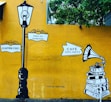 A mural with a vintage street lamp and signposts reading '10, Suffren Street' and '9:00am-7pm Tuesday Closed'. To the right, there is an illustration of a gramophone on top of a stack of books with the sign 'Café Des Arts'. The background is a bright yellow wall with green foliage at the top.