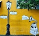 A mural with a vintage street lamp and signposts reading '10, Suffren Street' and '9:00am-7pm Tuesday Closed'. To the right, there is an illustration of a gramophone on top of a stack of books with the sign 'Café Des Arts'. The background is a bright yellow wall with green foliage at the top.