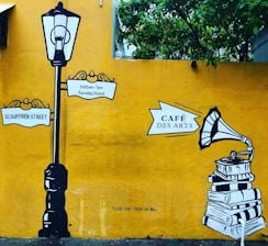A mural with a vintage street lamp and signposts reading '10, Suffren Street' and '9:00am-7pm Tuesday Closed'. To the right, there is an illustration of a gramophone on top of a stack of books with the sign 'Café Des Arts'. The background is a bright yellow wall with green foliage at the top.