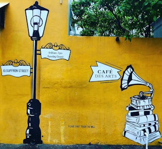 A mural with a vintage street lamp and signposts reading '10, Suffren Street' and '9:00am-7pm Tuesday Closed'. To the right, there is an illustration of a gramophone on top of a stack of books with the sign 'Café Des Arts'. The background is a bright yellow wall with green foliage at the top.