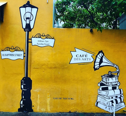 A mural with a vintage street lamp and signposts reading '10, Suffren Street' and '9:00am-7pm Tuesday Closed'. To the right, there is an illustration of a gramophone on top of a stack of books with the sign 'Café Des Arts'. The background is a bright yellow wall with green foliage at the top.