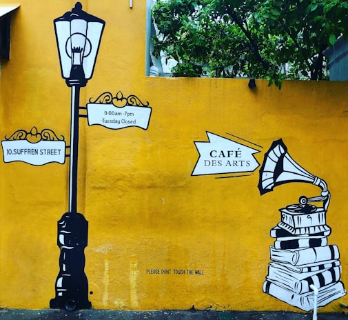 A mural with a vintage street lamp and signposts reading '10, Suffren Street' and '9:00am-7pm Tuesday Closed'. To the right, there is an illustration of a gramophone on top of a stack of books with the sign 'Café Des Arts'. The background is a bright yellow wall with green foliage at the top.