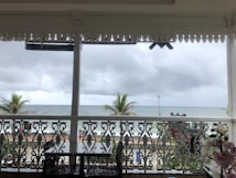 An ocean view from a covered terrace, featuring ornate railing and a black rectangular table with chairs. Two palm trees stand between the terrace and a distant view of the beach and the sea under a cloudy sky.