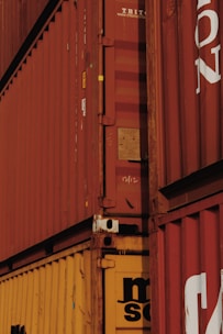 A close-up view of stacked shipping containers with red and yellow colors, featuring visible text and numbers on their surfaces. The containers have a rugged, industrial appearance with some signs of wear and rust around the edges.