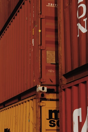 A close-up view of stacked shipping containers with red and yellow colors, featuring visible text and numbers on their surfaces. The containers have a rugged, industrial appearance with some signs of wear and rust around the edges.