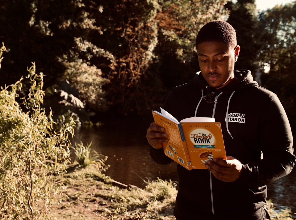 man in black jacket holding book near lake during daytime