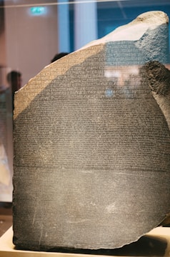 A large stone slab with inscriptions etched into it, showcasing multiple lines of text in different scripts. The surface appears worn with a rough and uneven edge, suggesting an ancient origin. The stone is displayed in a well-lit environment, possibly a museum, with reflections visible on its surface.