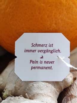 A close-up of a white tag with maroon-colored text that reads 'Schmerz ist immer vergänglich. Pain is never permanent.' The tag is attached to a string and placed against a background of ginger root and an orange.
