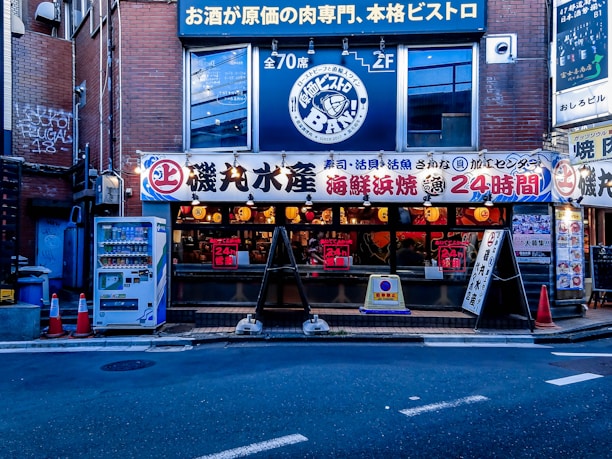 A street view of a Japanese restaurant with a brightly lit facade displaying various signs in Japanese. The establishment appears to be open 24 hours, and the signage suggests it specializes in seafood and bistro cuisine. Several orange traffic cones are placed on the sidewalk near a vending machine offering drinks.