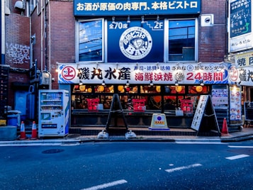 A street view of a Japanese restaurant with a brightly lit facade displaying various signs in Japanese. The establishment appears to be open 24 hours, and the signage suggests it specializes in seafood and bistro cuisine. Several orange traffic cones are placed on the sidewalk near a vending machine offering drinks.