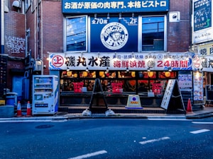 A street view of a Japanese restaurant with a brightly lit facade displaying various signs in Japanese. The establishment appears to be open 24 hours, and the signage suggests it specializes in seafood and bistro cuisine. Several orange traffic cones are placed on the sidewalk near a vending machine offering drinks.