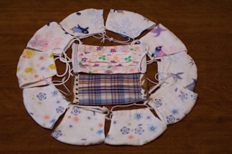 A collection of colorful, patterned face masks arranged in a circular pattern on a wooden surface. The masks feature various designs such as floral patterns, plaid, penguins, and abstract motifs.