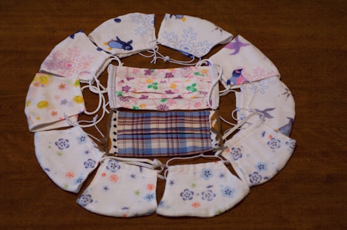 A collection of colorful, patterned face masks arranged in a circular pattern on a wooden surface. The masks feature various designs such as floral patterns, plaid, penguins, and abstract motifs.
