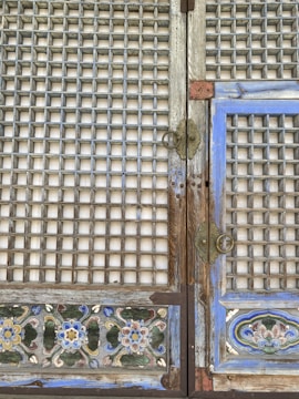 An intricately designed wooden door with a grid pattern, adorned with colorful floral motifs at the bottom. The wood appears weathered, with a faded light blue hue outlining the panels, and brass handles and hinges provide an antique look.