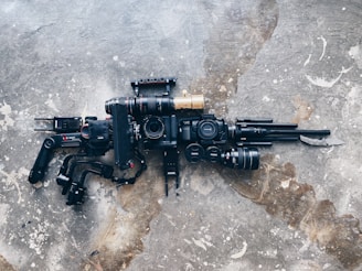 A collection of camera equipment is laid out on a textured concrete surface. The gear includes several camera bodies, lenses, and stabilizing equipment arranged to form a shape that resembles a rifle.
