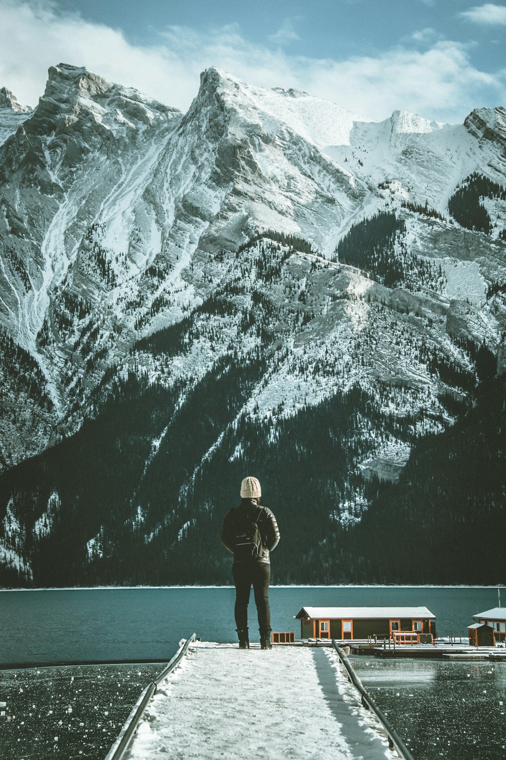 man in black jacket and black pants standing on white bench looking at snow covered mountain
