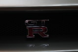 The image features a close-up view of a GT-R emblem on the grille of a car. The emblem consists of metallic letters with a red 'R' highlighted against a dark mesh background.