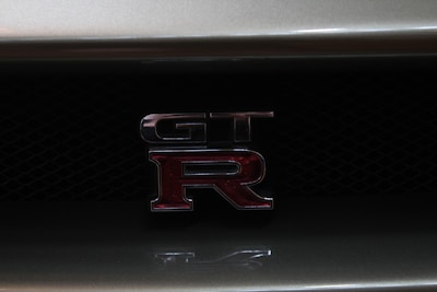 The image features a close-up view of a GT-R emblem on the grille of a car. The emblem consists of metallic letters with a red 'R' highlighted against a dark mesh background.