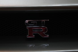 The image features a close-up view of a GT-R emblem on the grille of a car. The emblem consists of metallic letters with a red 'R' highlighted against a dark mesh background.