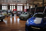 A car exhibition is taking place in a spacious, well-lit indoor setting. Several vintage and modern cars are on display, including a blue sports car in the foreground and a green vintage car on the left side. People are walking around and observing the vehicles. There are large windows, high ceilings, and a series of display panels on the walls.