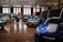 A car exhibition is taking place in a spacious, well-lit indoor setting. Several vintage and modern cars are on display, including a blue sports car in the foreground and a green vintage car on the left side. People are walking around and observing the vehicles. There are large windows, high ceilings, and a series of display panels on the walls.