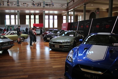 A car exhibition is taking place in a spacious, well-lit indoor setting. Several vintage and modern cars are on display, including a blue sports car in the foreground and a green vintage car on the left side. People are walking around and observing the vehicles. There are large windows, high ceilings, and a series of display panels on the walls.
