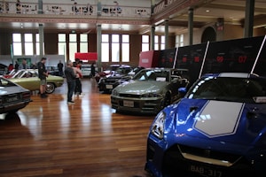A car exhibition is taking place in a spacious, well-lit indoor setting. Several vintage and modern cars are on display, including a blue sports car in the foreground and a green vintage car on the left side. People are walking around and observing the vehicles. There are large windows, high ceilings, and a series of display panels on the walls.