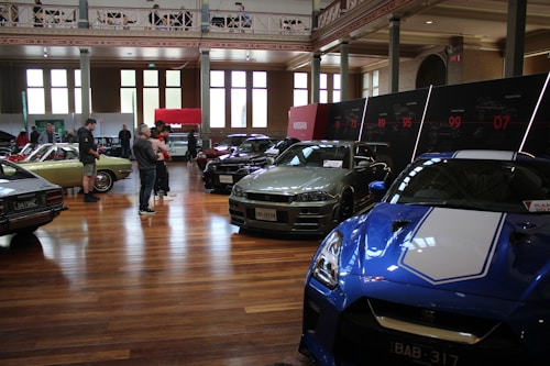 A car exhibition is taking place in a spacious, well-lit indoor setting. Several vintage and modern cars are on display, including a blue sports car in the foreground and a green vintage car on the left side. People are walking around and observing the vehicles. There are large windows, high ceilings, and a series of display panels on the walls.