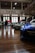 A car exhibition inside a historic building with polished wooden floors and high ceilings. Several modern and classic cars are lined up, showcasing their designs. People are walking around, observing the cars and interacting with each other. There is a visible balcony with more attendees looking down at the display.