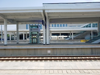 A modern train station platform with multiple tracks and an accessible elevator. The platform is clean and well-structured with overhead roofing. A high-speed train labeled CRH is partially visible in the background. There are also ramps and stairs leading to elevated sections.