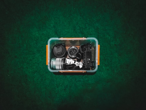 A storage container placed on a green surface holds various camera equipment. Inside the container, there are camera lenses, a camera body, and other photography accessories. The container has orange handles on either side.