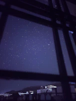 A night sky with twinkling stars seen through a window framed by sheer curtains.
