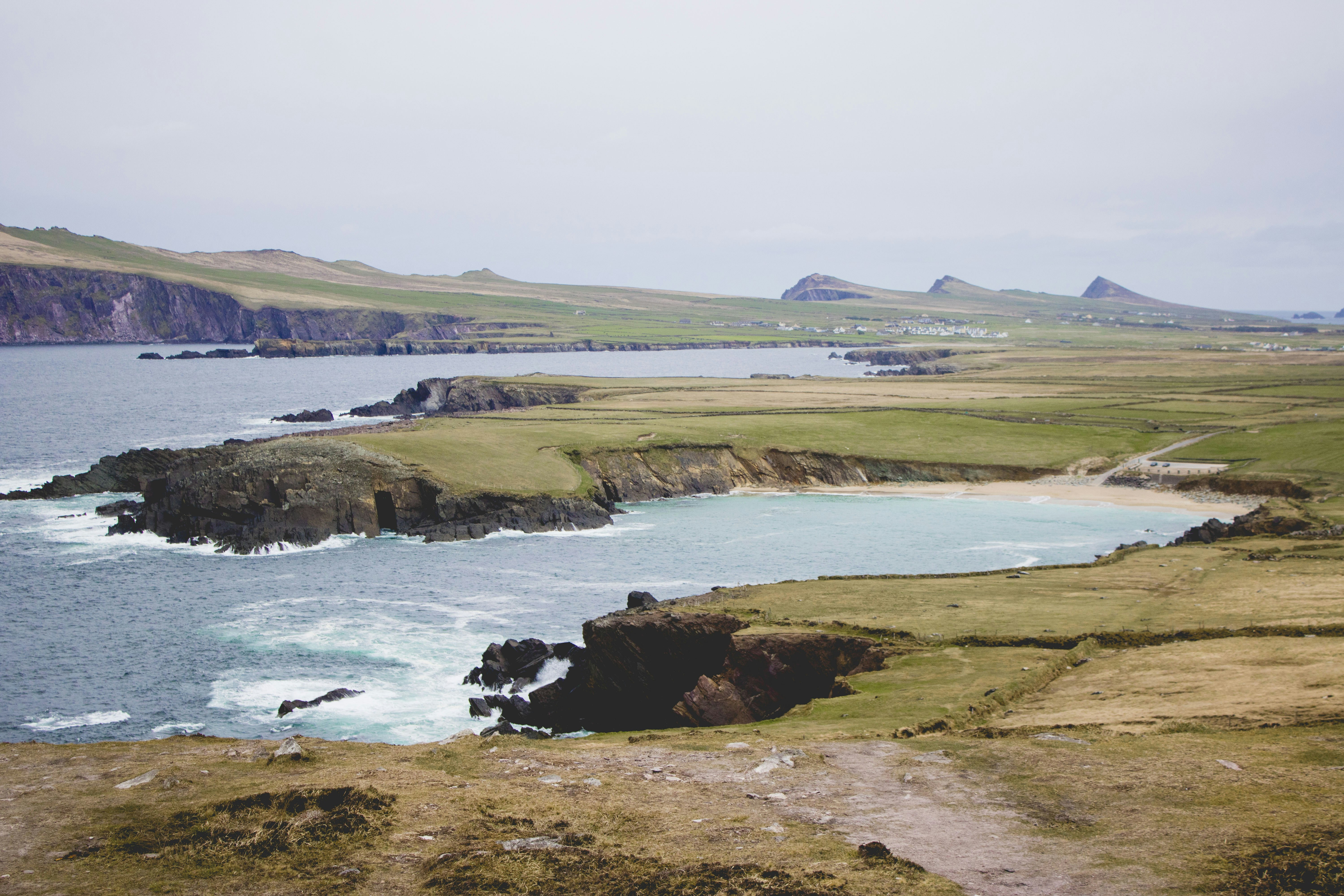 green grass field near body of water during daytime, A bay on Slea Head Drive with three peaks in the background known as "The Tree Sisters"