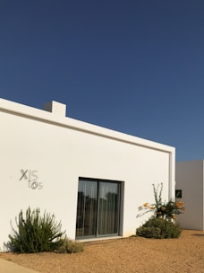 A minimalist white building with a modern, flat roof is set against a clear blue sky. The facade features a set of glass sliding doors and is accented with some green shrubbery and yellow flowers in the landscaping. The word 'Xistos' is displayed on the wall.