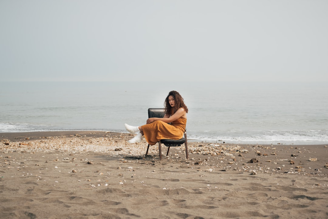 woman in brown jacket and white pants sitting on brown wooden chair on beach during daytime