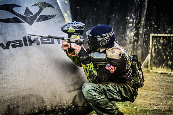 A person wearing protective gear and a mask is crouched behind a large inflatable obstacle, holding a paintball marker. The outfit is adorned with various patches and an American flag, suggesting participation in a competitive event. The background appears to be a paintball arena with splattered paint visible on surfaces.