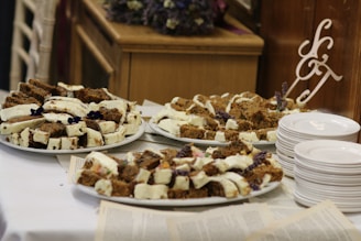 Several plates are filled with slices of cake topped with a white frosting and decorated with purple flowers. The cakes are arranged on white platters placed on a table covered with what seems to be pages or papers. Beside the plates is a stack of white ceramic dishes and a decorative monogram with letters. The setting has a rustic and cozy feel, with wooden furniture visible in the background.
