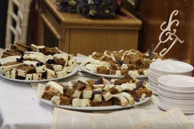 Several plates are filled with slices of cake topped with a white frosting and decorated with purple flowers. The cakes are arranged on white platters placed on a table covered with what seems to be pages or papers. Beside the plates is a stack of white ceramic dishes and a decorative monogram with letters. The setting has a rustic and cozy feel, with wooden furniture visible in the background.