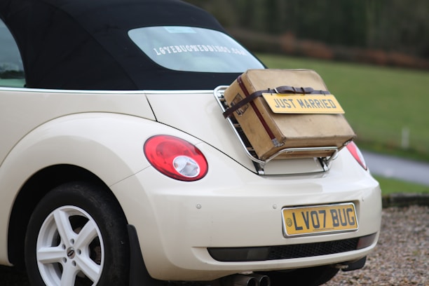 A cream-colored convertible car with a black soft top has a vintage suitcase strapped to its rear, featuring a 'Just Married' sign. The vehicle's license plate reads 'LV07 BUG'. The scene is set on a gravel road by a green field.