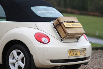 A cream-colored convertible car with a black soft top has a vintage suitcase strapped to its rear, featuring a 'Just Married' sign. The vehicle's license plate reads 'LV07 BUG'. The scene is set on a gravel road by a green field.