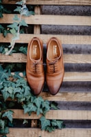 A pair of polished brown leather dress shoes is placed on a wooden pallet, surrounded by lush green ivy leaves. The shoes are neatly laced and positioned side by side, showcasing their craftsmanship. The background consists of wooden planks with a rustic finish, adding a natural and earthy feel to the scene.