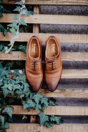 A pair of polished brown leather dress shoes is placed on a wooden pallet, surrounded by lush green ivy leaves. The shoes are neatly laced and positioned side by side, showcasing their craftsmanship. The background consists of wooden planks with a rustic finish, adding a natural and earthy feel to the scene.