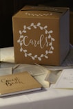 A cardboard box labeled 'Cards' is placed on a table. The box is decorated with a white floral wreath design. Surrounding the box are scattered pages from books and a guest book, which is closed and sits nearby.