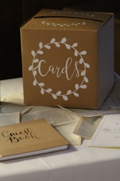 A cardboard box labeled 'Cards' is placed on a table. The box is decorated with a white floral wreath design. Surrounding the box are scattered pages from books and a guest book, which is closed and sits nearby.